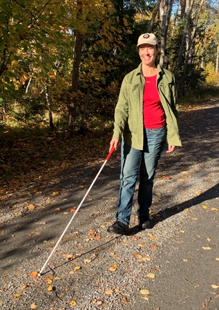 Leende kvinna i jeans och keps går på en grusväg med den vita käppen i höger hand.