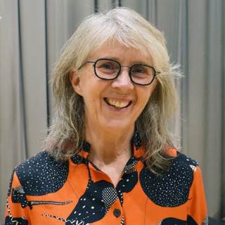 Inger Berndtsson har grått hår ner till axlarna och glasögon med svarta skalmar. Hon bär en mönstrad blus i svart och orange.